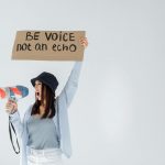 Empowered woman shouts message through megaphone, holding sign that reads 'Be voice, not an echo'.